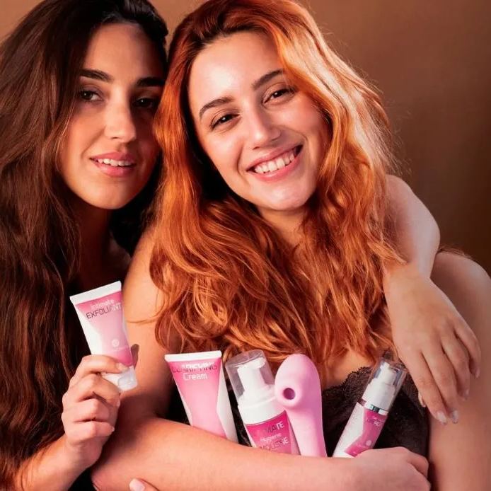 Two adult women smiling while holding a selection of pink Femintimate intimate care products, including tubes and bottles, in a studio setting.