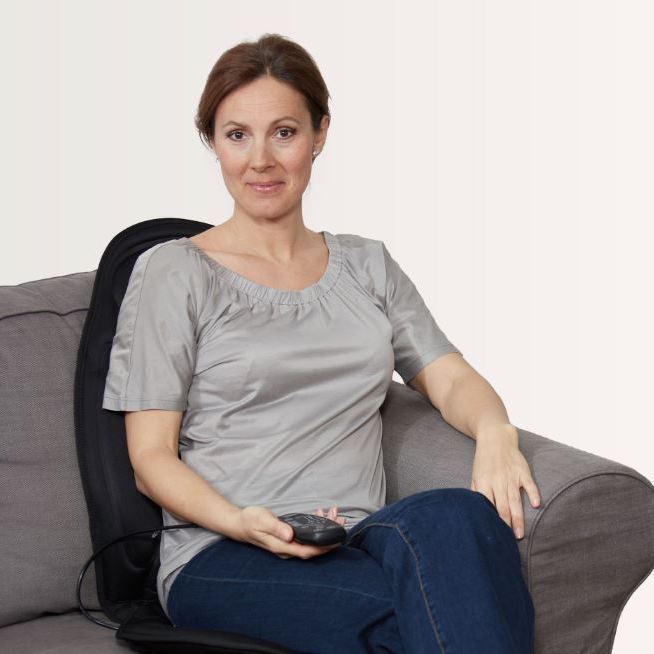 Woman sitting on a sofa using a Lanaform remote-controlled vibrating heated back massager cushion, providing targeted back pain relief and muscle relaxation at home with adjustable massage zones and heat.