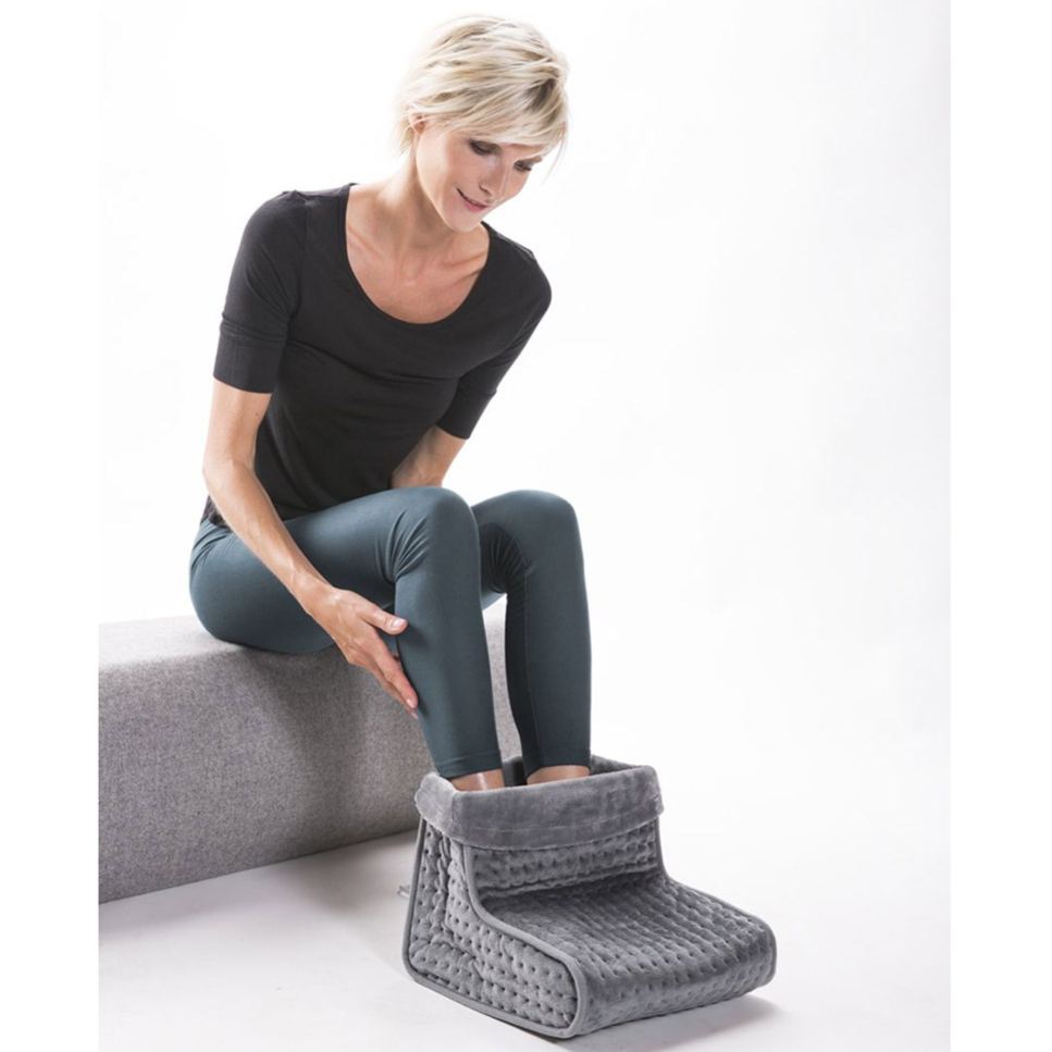 Woman sitting on a seat using a grey Lanaform electric heated foot warmer, gently warming her feet to improve comfort and circulation indoors, ideal for relieving cold feet during rest or relaxation.