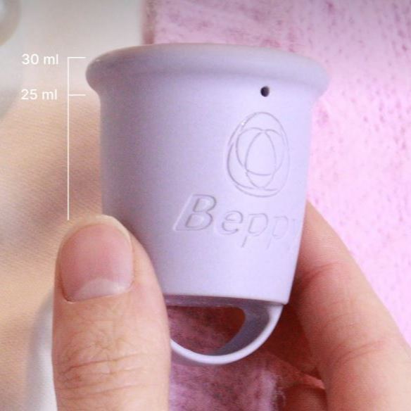 Close-up of a Beppy reusable menstrual cup held between fingers, showing the cup shape, embossed Beppy logo, loop handle, and printed measurement markings on the side.