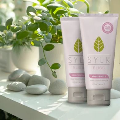 Two SYLK BLISS natural water-based lubricant tubes displayed beside green plants and smooth stones, showcasing the plant-based, pH-balanced new formula designed to support gentle intimate comfort, hydration, and a natural, calming experience.