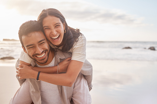 Smiling couple at the beach with the woman hugging the man from behind as they laugh together, standing near the shoreline on a bright, sunny day. The Life-Changing Device That All Men Should Know About-StressNoMore