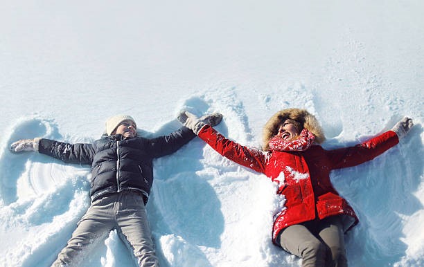 Two people lying in the snow making snow angels, dressed in winter coats and holding hands while enjoying a snowy outdoor scene. Is It My Imagination, Or Do I Pee More During Winter?-StressNoMore