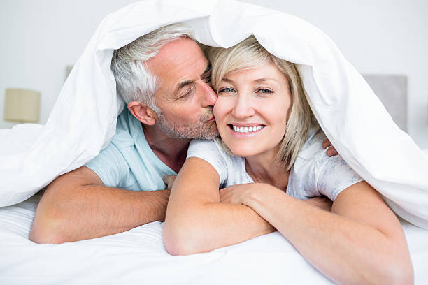 The Truth about Sex and the Pelvic Floor Over 40-StressNoMore. Happy older couple cuddling under a duvet, with the man kissing the woman’s cheek, symbolising intimacy, connection, healthy relationships and midlife wellbeing.