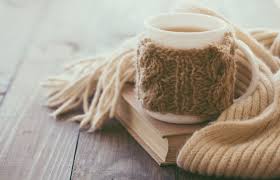 Warm drink in a mug wrapped in a knitted cosy, placed on top of a book with a soft scarf beside it on a wooden surface. Winter Wellness: Comprehensive Solutions for Seasonal Ailments. Stressnomore