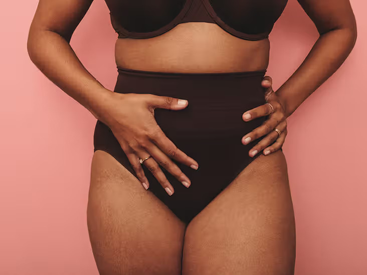 Contiform: Unique Vaginal Pessary Available on NHS-StressNoMore. woman in dark high-waisted underwear touching her lower abdomen while standing against a pink background.