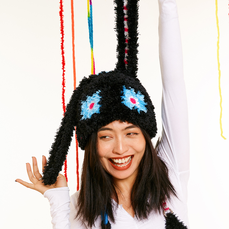 Playful front view of a model holding up one long ear of the black Bunny Ear Crochet Hat, showing the vibrant blue flower details.