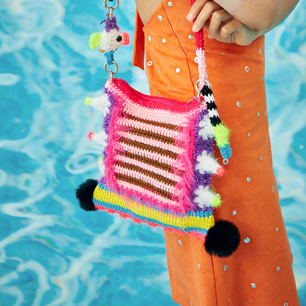 Side lifestyle view of a model holding the striped side of the colorful crochet shoulder bag, highlighting the multi-toned long strap with small cute ornaments.