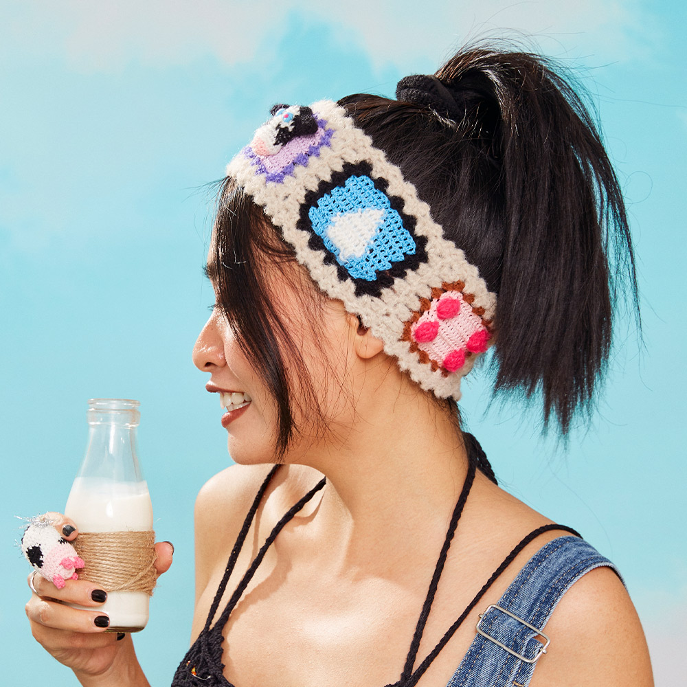 Side profile view of a model holding a milk bottle, showcasing the vibrant blue and pink crochet block patterns on the fluffy velvet headband.