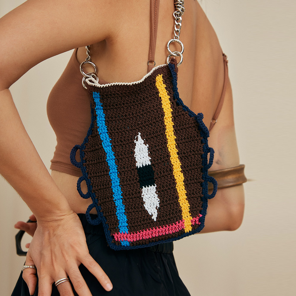 A model styling the eye color matching vase crochet crossbody bag at her waist, showcasing the bright blue and yellow vertical stitch stripes against a brown top.