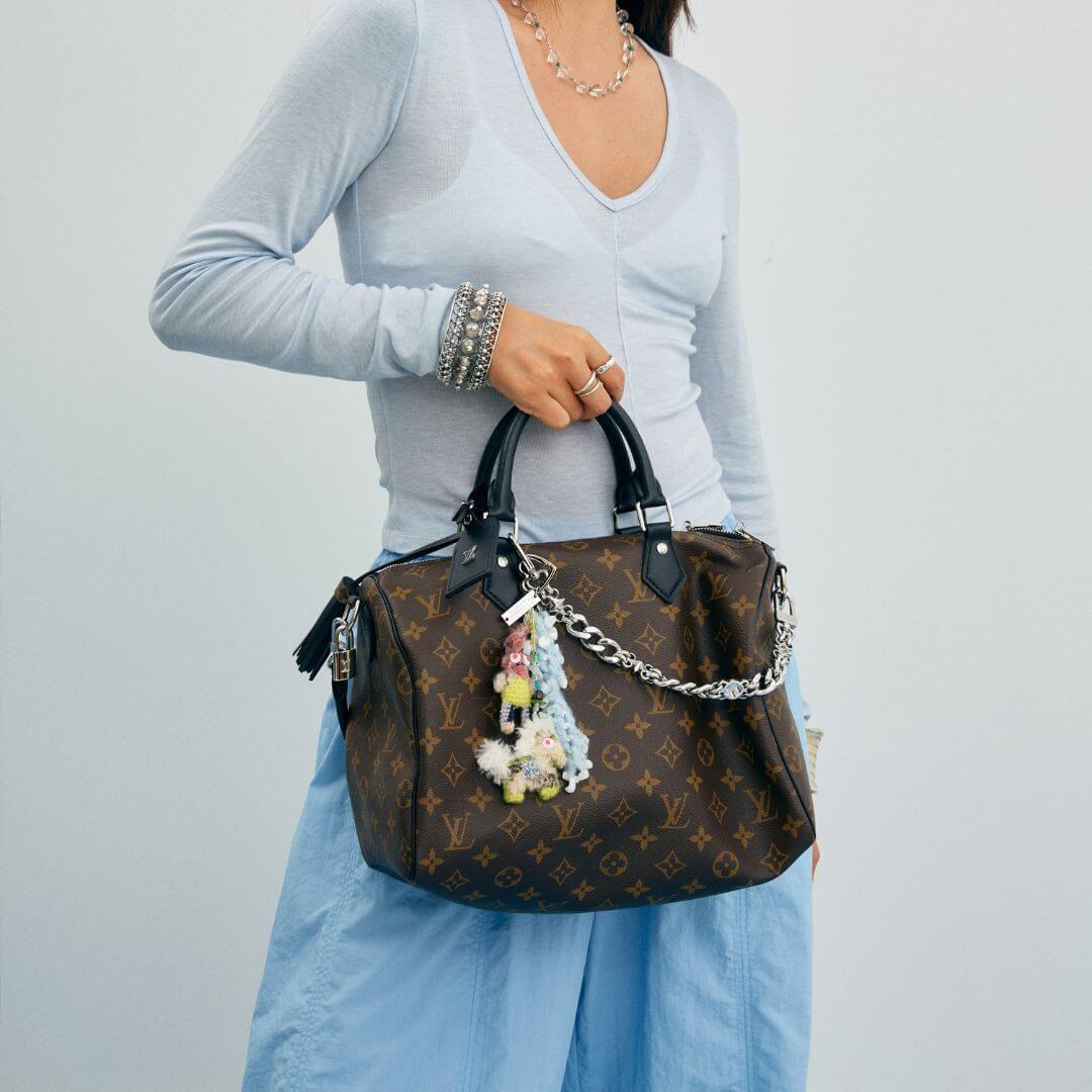 A lifestyle shot demonstrating the Feeky crochet charm attached to the handle of a luxury patterned handbag, worn with a light blue top.