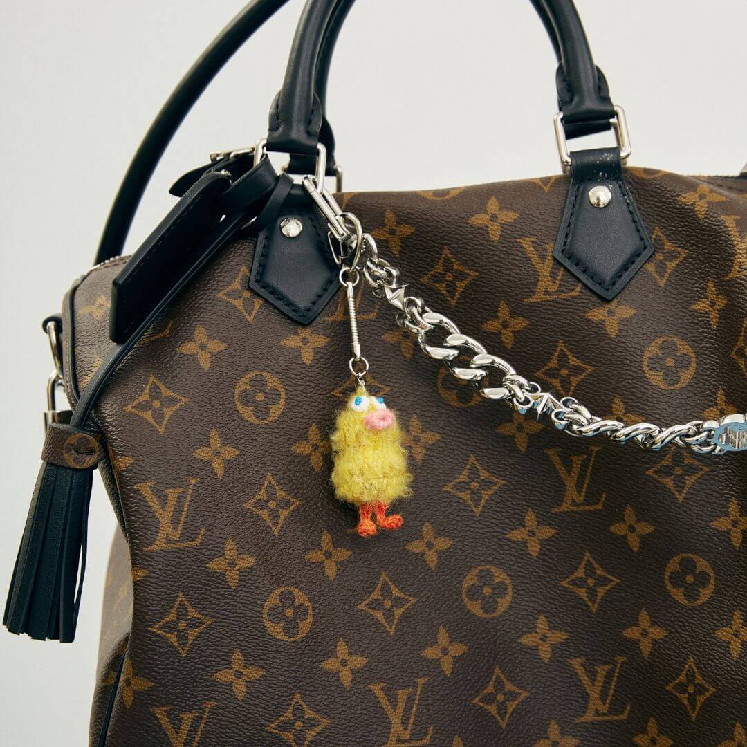 A lifestyle shot of the yellow Ugly Duckling charm attached to a luxury patterned handbag, demonstrating its whimsical styling potential.
