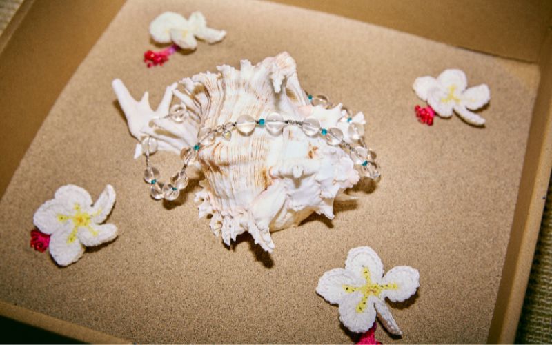 {"default":"Delicate white crochet jewelry displayed on a large seashell with tropical flowers, reflecting the wearable fairytale brand concept."}
