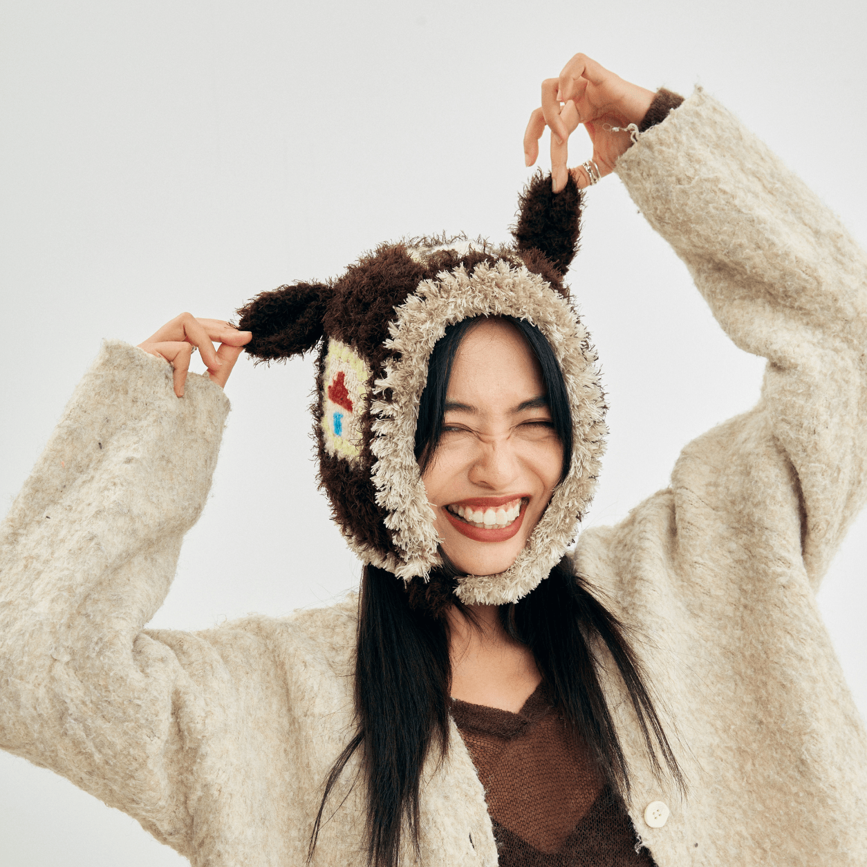 Smiling woman playfully holding the ears of her Little Lamb Patchwork Hat while wearing a cozy cardigan.