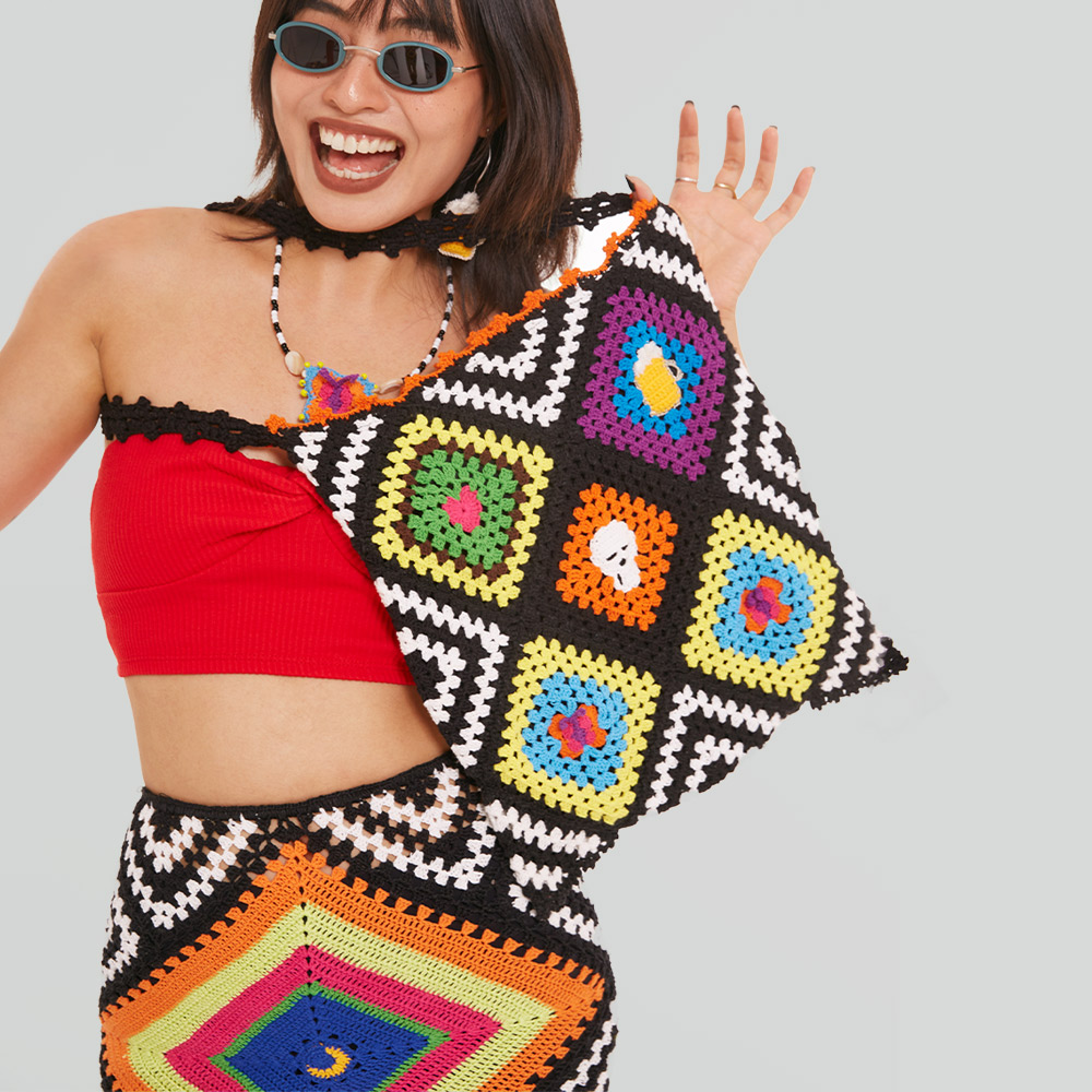 A joyful model wearing sunglasses holding up the Patchwork Crochet Tote Bag, showcasing the highly saturated floral stitch blocks and soft heavy yarn texture.