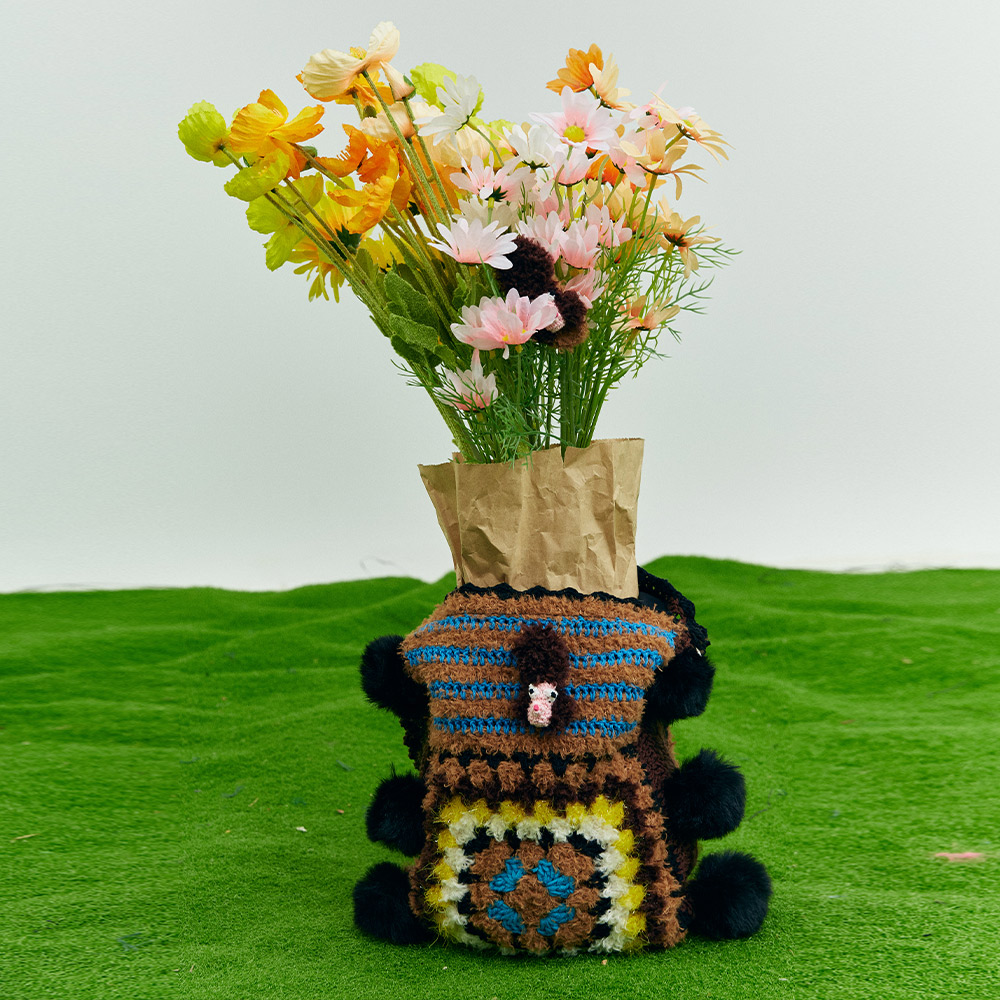 The puppy theme crochet bag used creatively as a decorative sleeve for a flower bouquet on green grass, displaying an artisan lifestyle aesthetic.