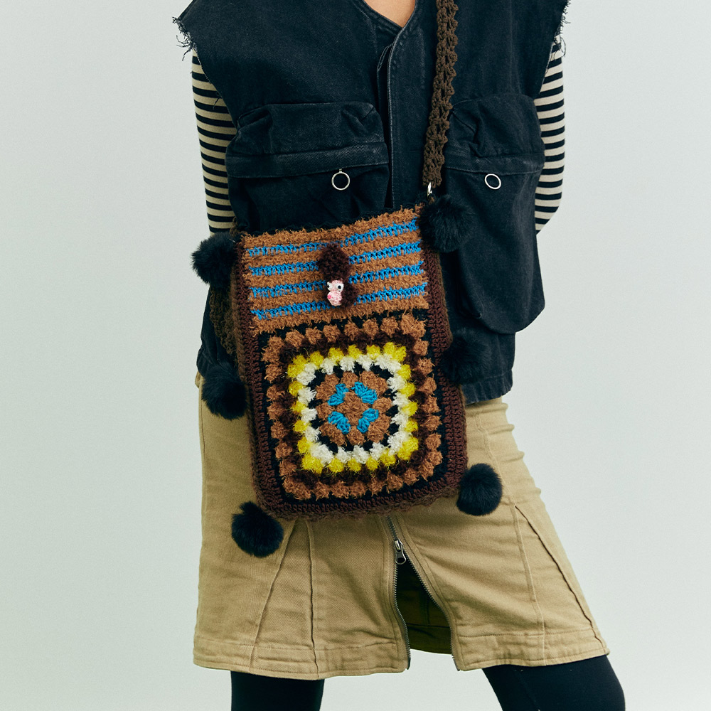 Side profile view of a model carrying the Puppy Theme Crochet Crossbody Bag on green grass, showcasing the colorful center granny square and attached fuzzy pom-poms.