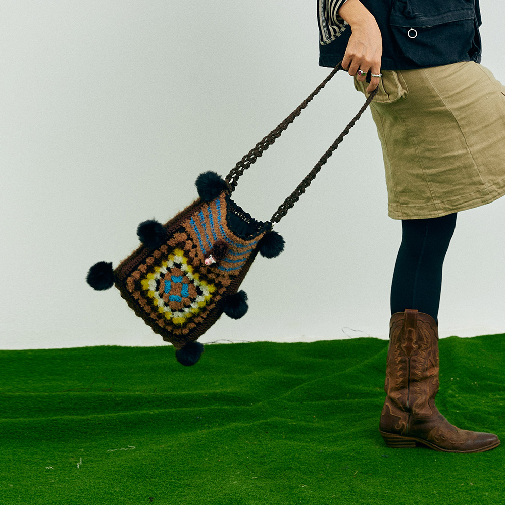 A model holding the long strap of the puppy theme crochet shoulder bag downwards, paired with retro brown cowboy boots for an autumn western look.