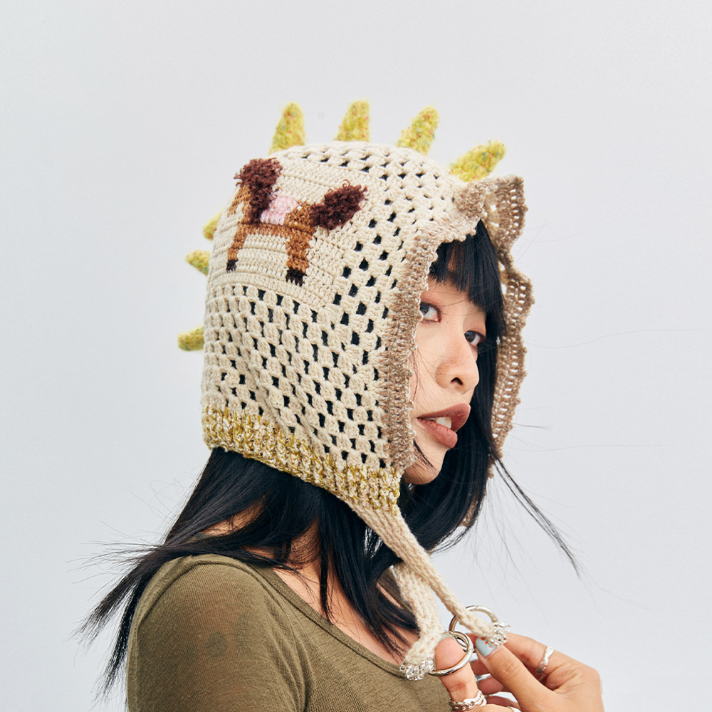 An angled three-quarter view of a model wearing the beige Unicorn Pony Crochet hood hat with dangling earflap ties.