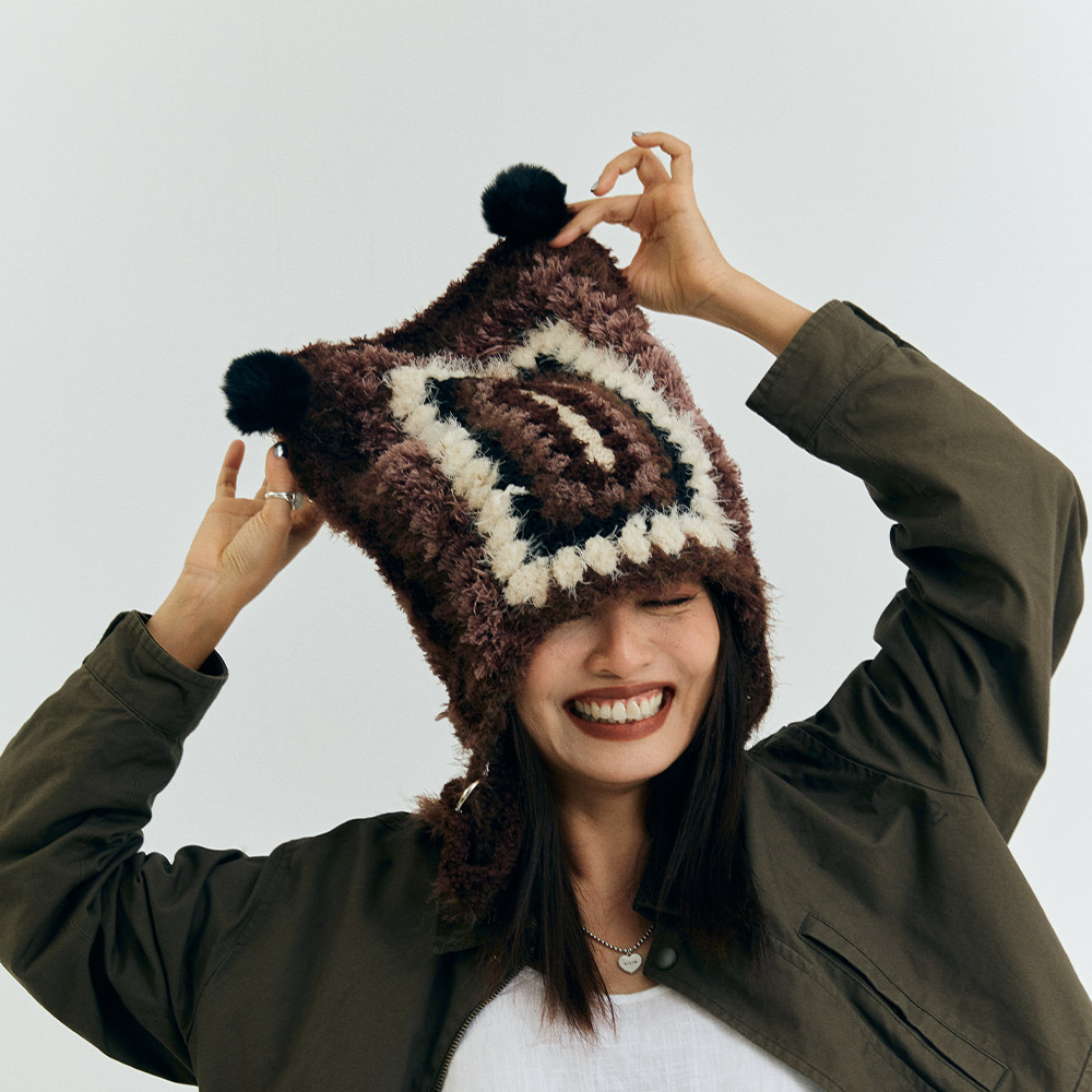 A smiling model wearing the 2-in-1 product transformable into a vintage grid crochet cat ear hat with playful black fuzzy poms.