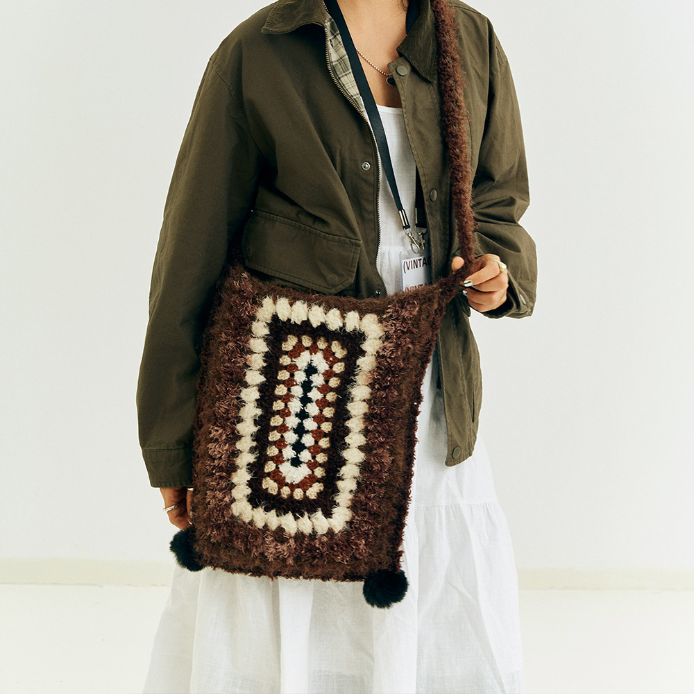 Front view of a model wearing the Vintage Grid Crochet Crossbody Bag, showcasing the concentric brown and cream geometric stitch pattern and black pom-pom accents.