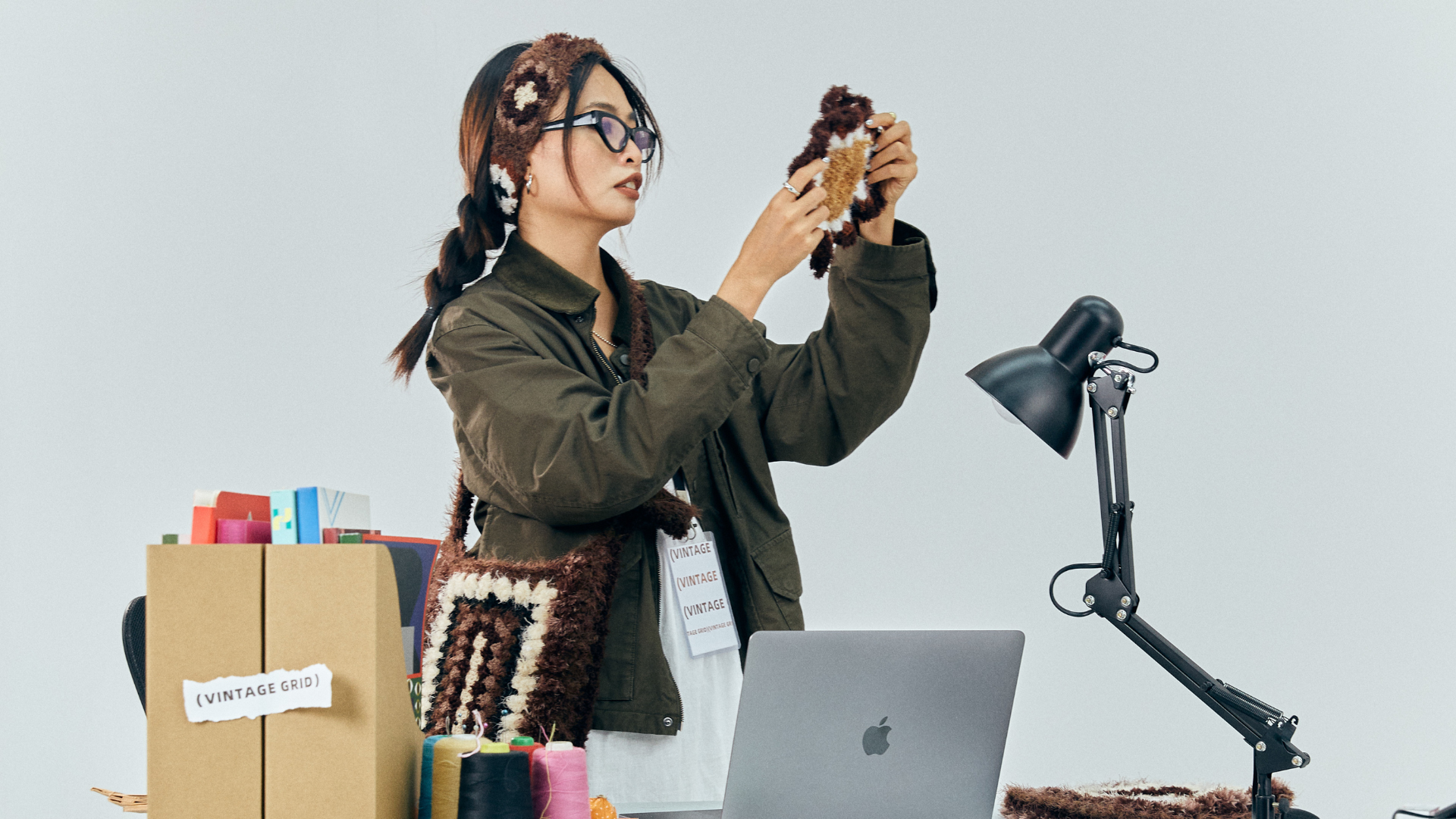 A female entrepreneur standing at a desk, carefully inspecting a handmade crochet item, with a laptop and a desk lamp, featuring vibrant dopamine style colors in the workspace.