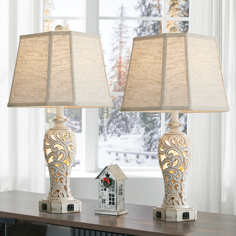 A pair of washed white table lamp with carved branch & leaf pattern, the lamps are sitting on a table in a living room.
