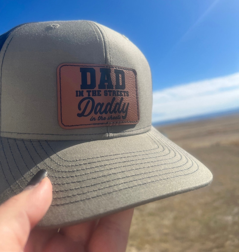 May include: A green and black baseball cap with a brown leather patch that says 'Dad in the streets Daddy in the sheets'.