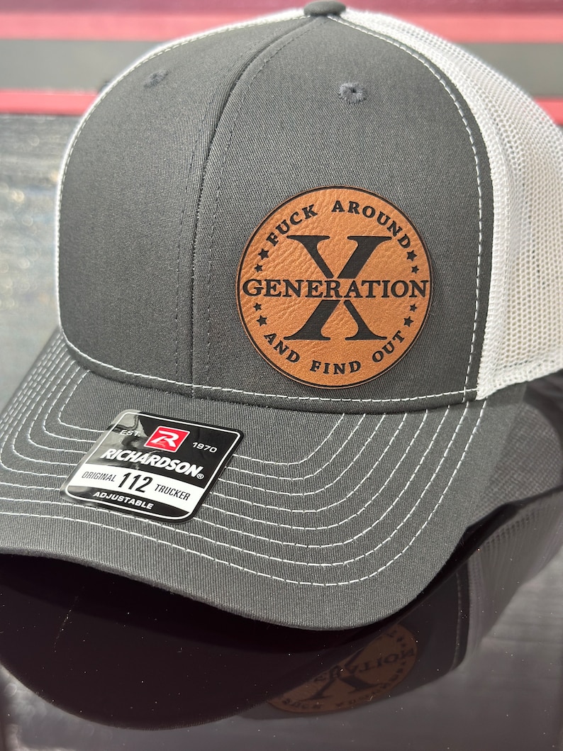 May include: Gray and white adjustable Richardson 112 trucker hat. Features a brown leather patch with the text 'FUCK AROUND GENERATION X AND FIND OUT' in a circular design. The hat is a classic style with a mesh back and curved brim.
