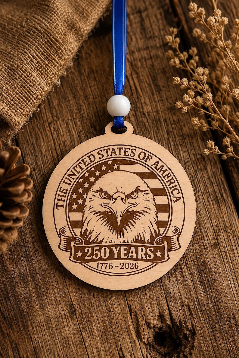 May include: A round wooden ornament with a patriotic design. It features an eagle, stars, and stripes, with the text "THE UNITED STATES OF AMERICA" and "250 YEARS 1776-2026". The ornament hangs from a blue ribbon.