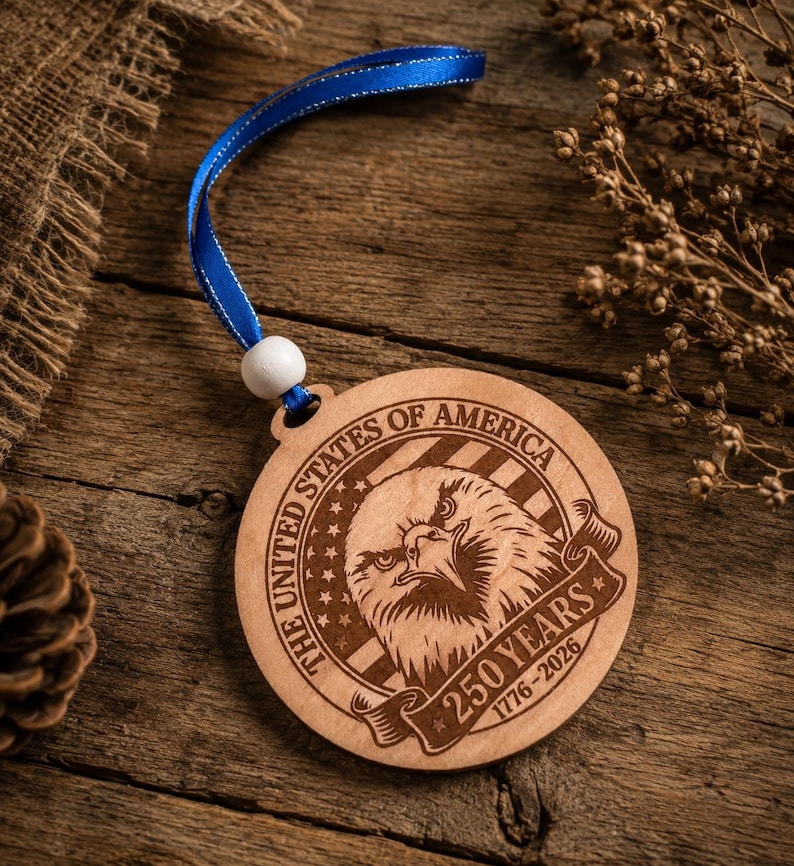 May include: A round wooden ornament with a blue ribbon. The ornament features an engraved eagle design with the text "THE UNITED STATES OF AMERICA" and "250 YEARS 1776-2026". The ornament is made of light-colored wood.