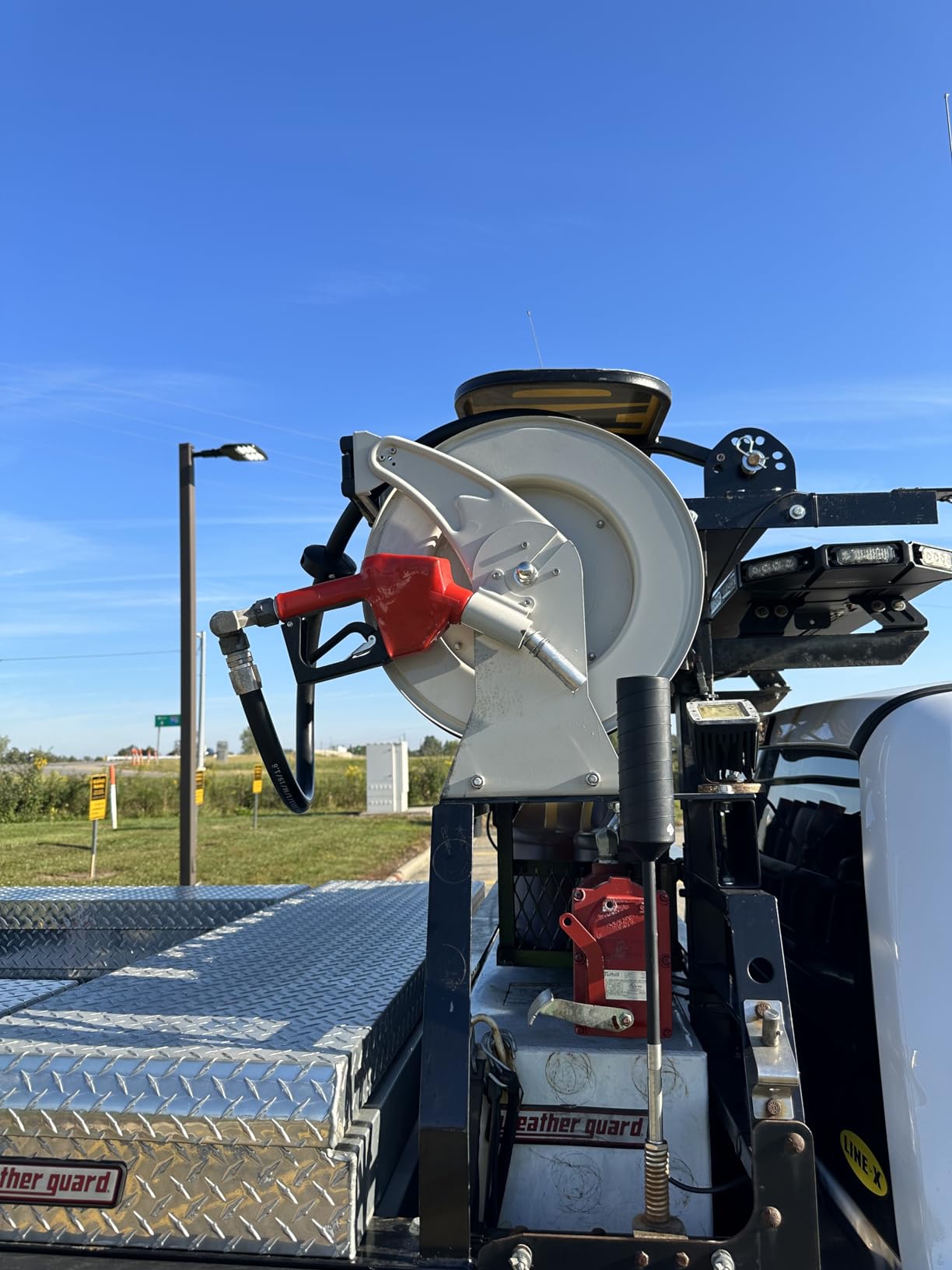 A heavy-duty fuel hose reel with a red nozzle mounted on the back of a truck, equipped with a durable metal frame and positioned on a diamond-patterned steel platform under a clear blue sky.