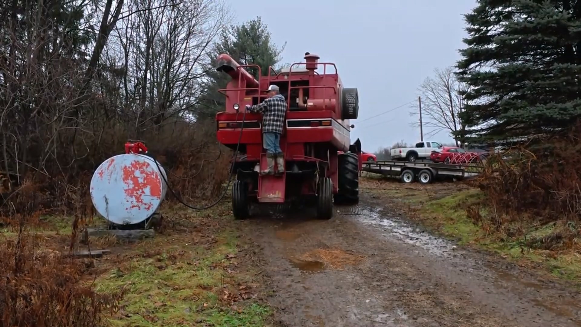 An agricultural worker refueling a large red harvester in a muddy rural field using a fuel transfer pump connected to a white storage tank. Trees and vehicles are visible in the background