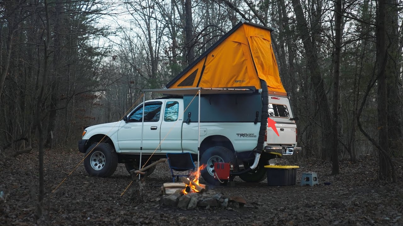 A white Toyota Tacoma with an orange rooftop tent and diesel heater in a forest camping setup.