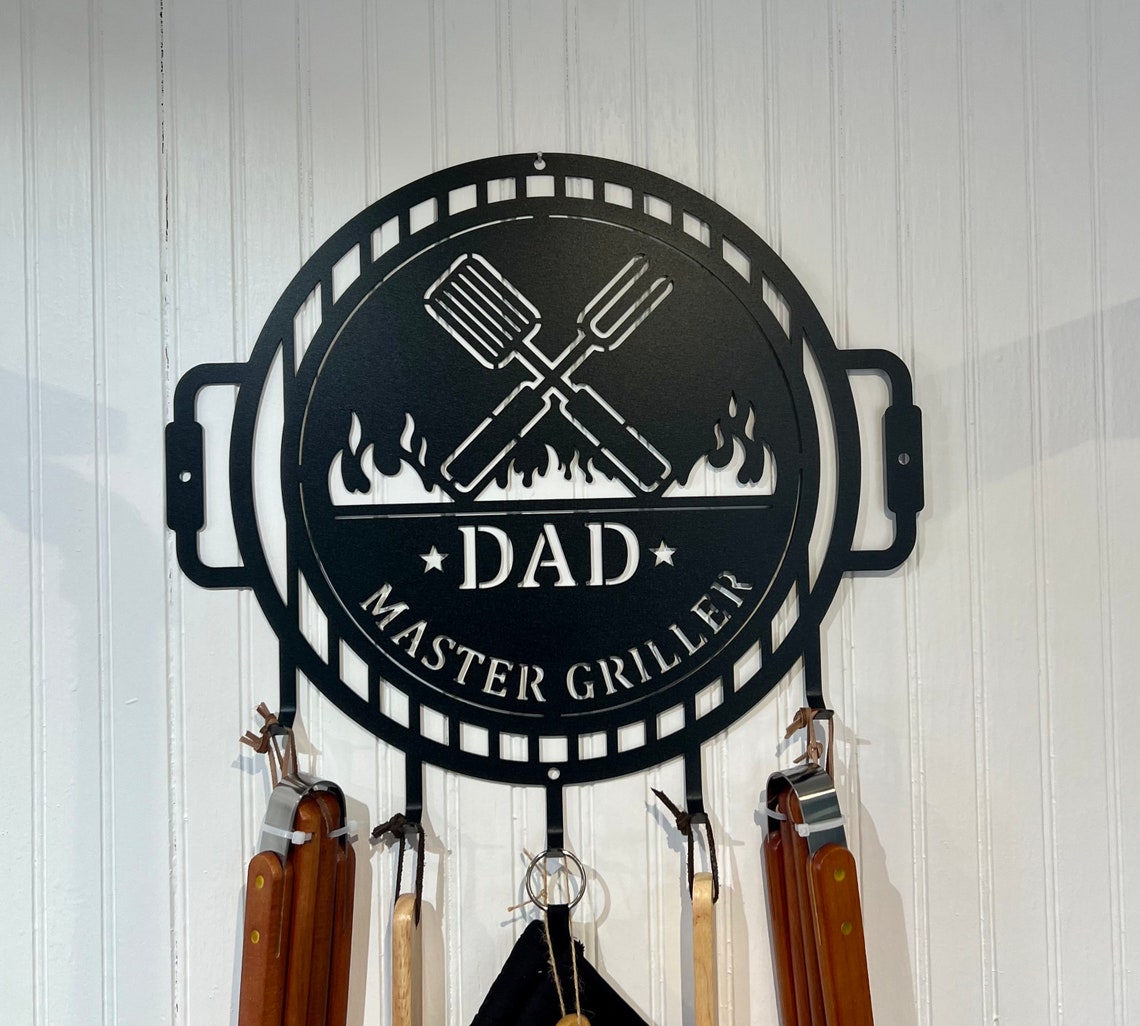 烧烤大师金属标牌，带黑色餐具挂架，白色木墙上显示个性化爸爸姓名文字，并悬挂烧烤器具