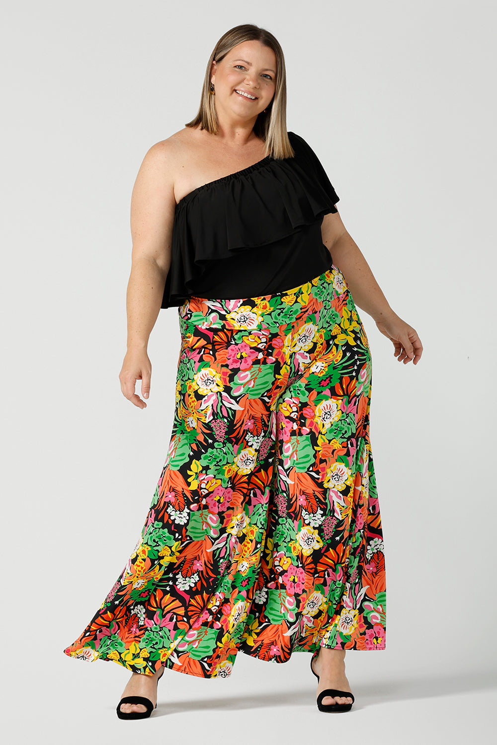 A curvy size 18 woman wears the Caspian Palazzo pant in a beautiful bright colourful print. In soft and lightweight jersey this pant features side pocket and a wide leg opening. Styled back with a black off shoulder top and black heels. Made in Australia 