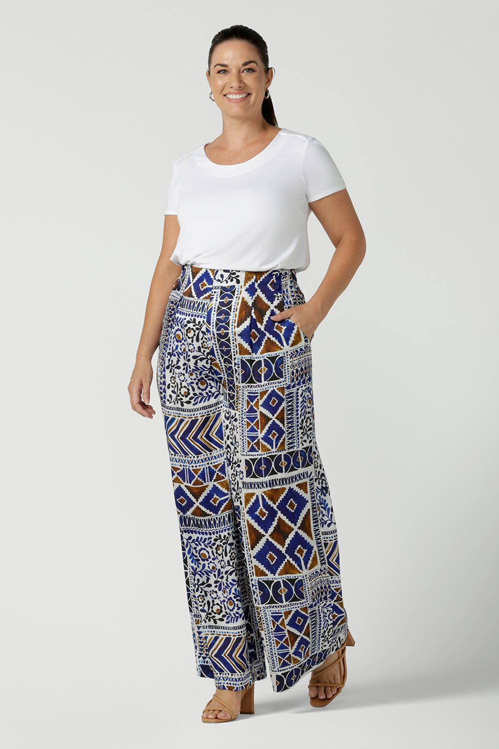 Size 12 woman wears a Ellery pant in soft Italian Viscose fabric. Digitally printed in a Cadaques tile print. A pleat front style with self covered buttons and a wide leg opening and functioning pockets. Women wears a high waist satin wide leg pants in a 