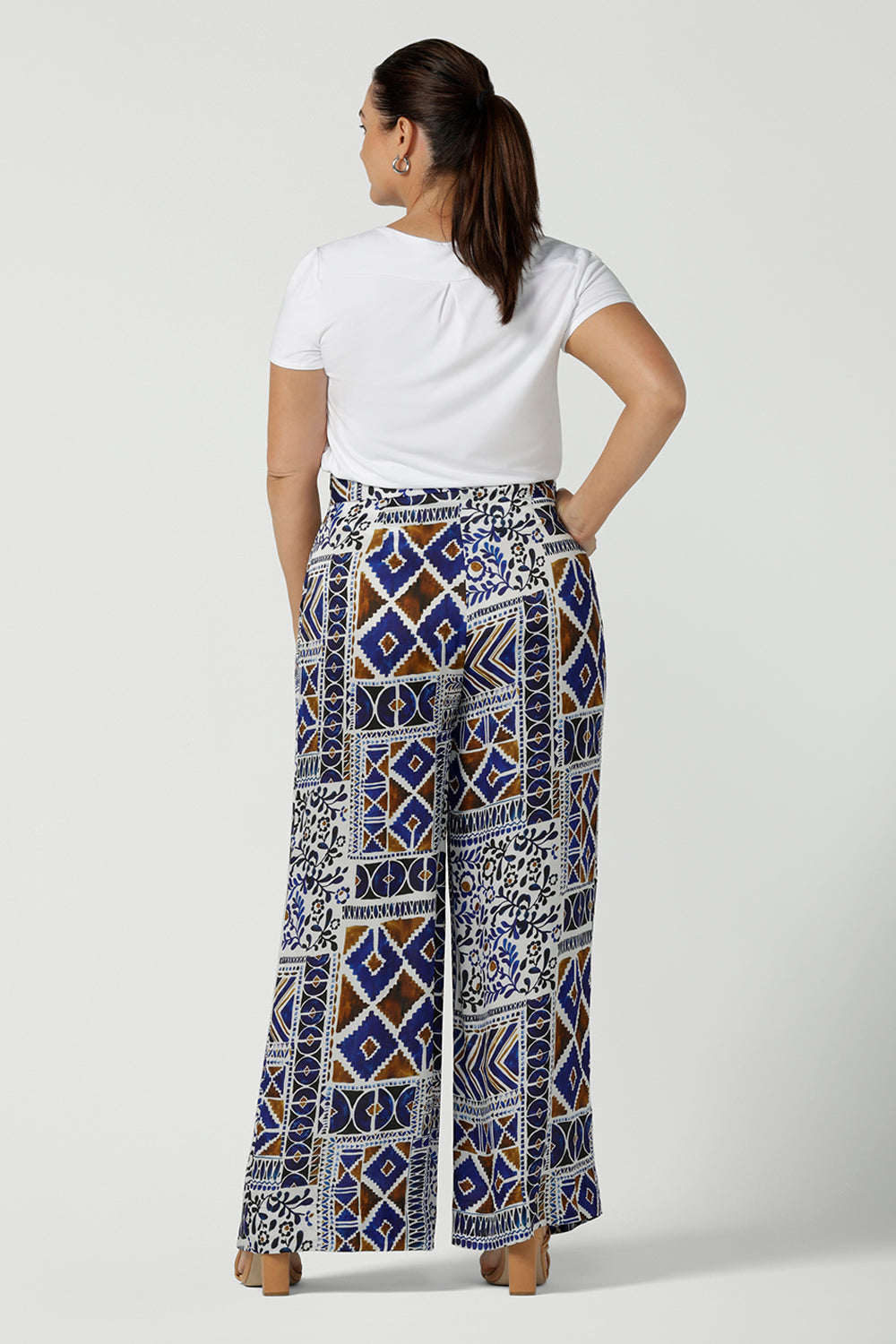 Back view of a size 12 woman wears a Ellery pant in soft Italian Viscose fabric. Digitally printed in a Cadaques tile print. A pleat front style with self covered buttons and a wide leg opening and functioning pockets. Women wears a high waist satin wide 