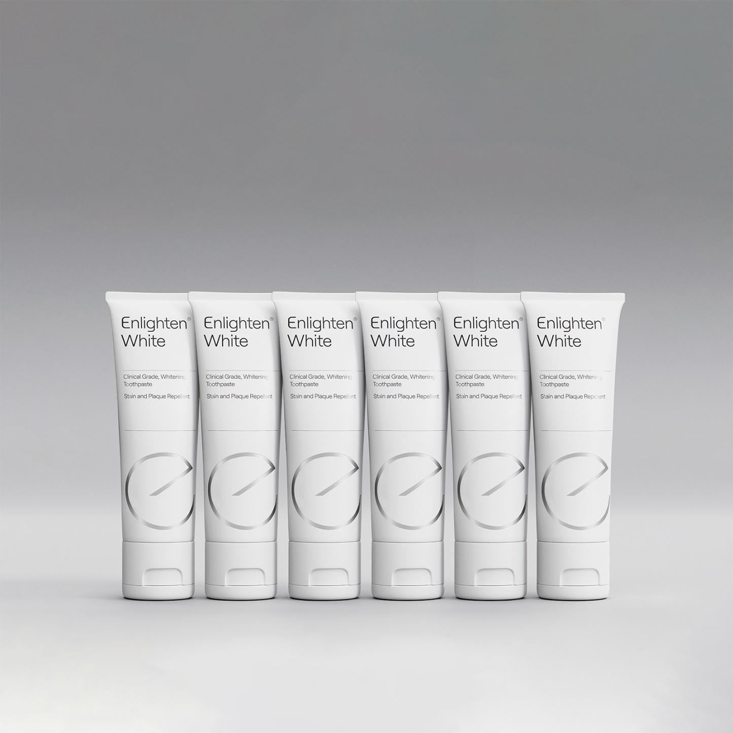 A row of six tubes of Enlighten White toothpaste displayed against a neutral grey background. Each tube is white with the Enlighten logo and branding, featuring the tagline "Clinical Grade, Whitening Toothpaste – Stain and Plaque Repellent." The clean and minimalist layout highlights the premium and professional nature of the product.