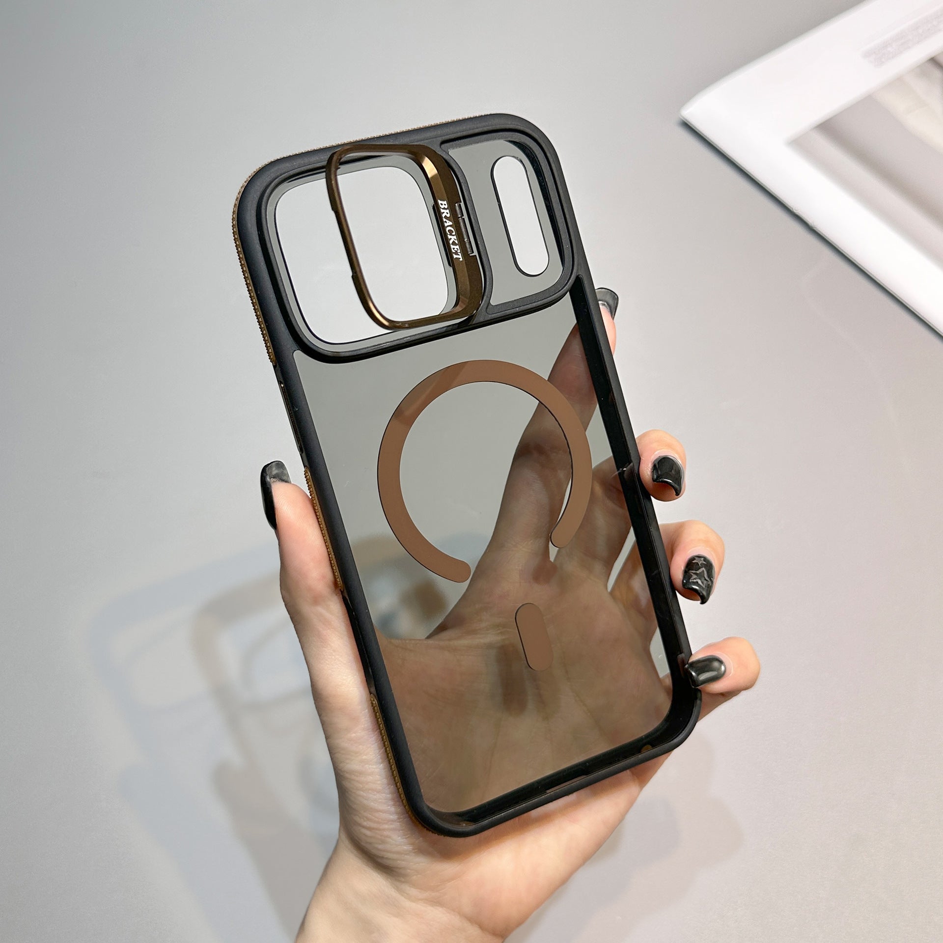 CaseSun-Hand displaying an iPhone 17 Pro Max case with a matte translucent back, bronze MagSafe ring, and raised lens bracket stand.