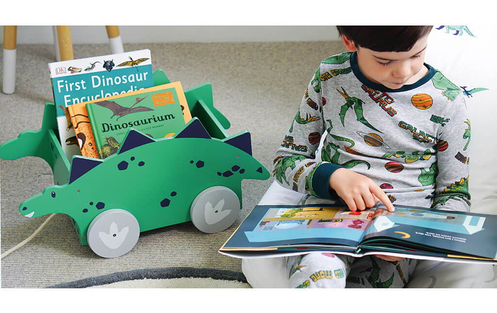 Young boy reading a book next to dinosaur book cart