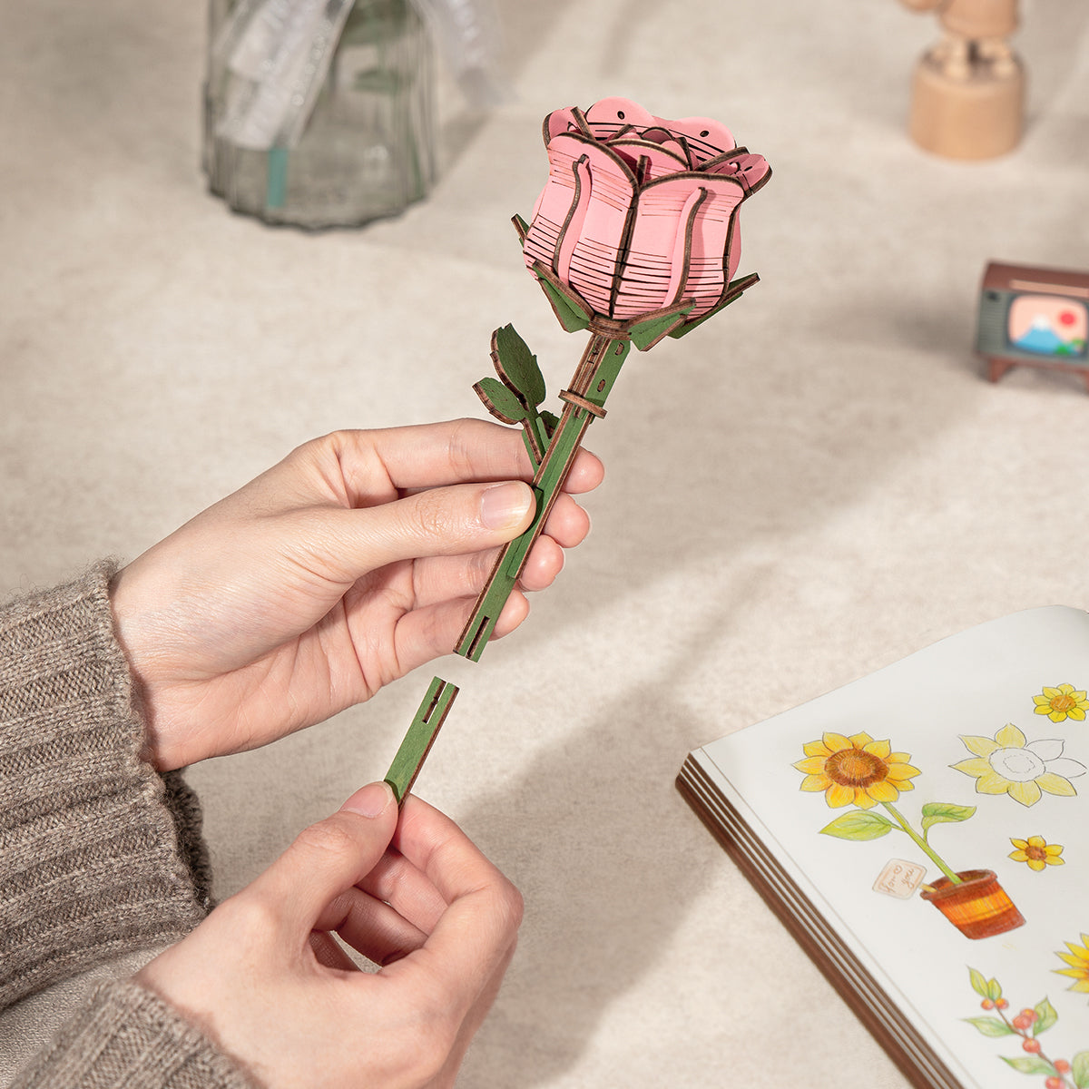 Rowood wooden pink rose stem is held by hand above a tabletop craft scene.