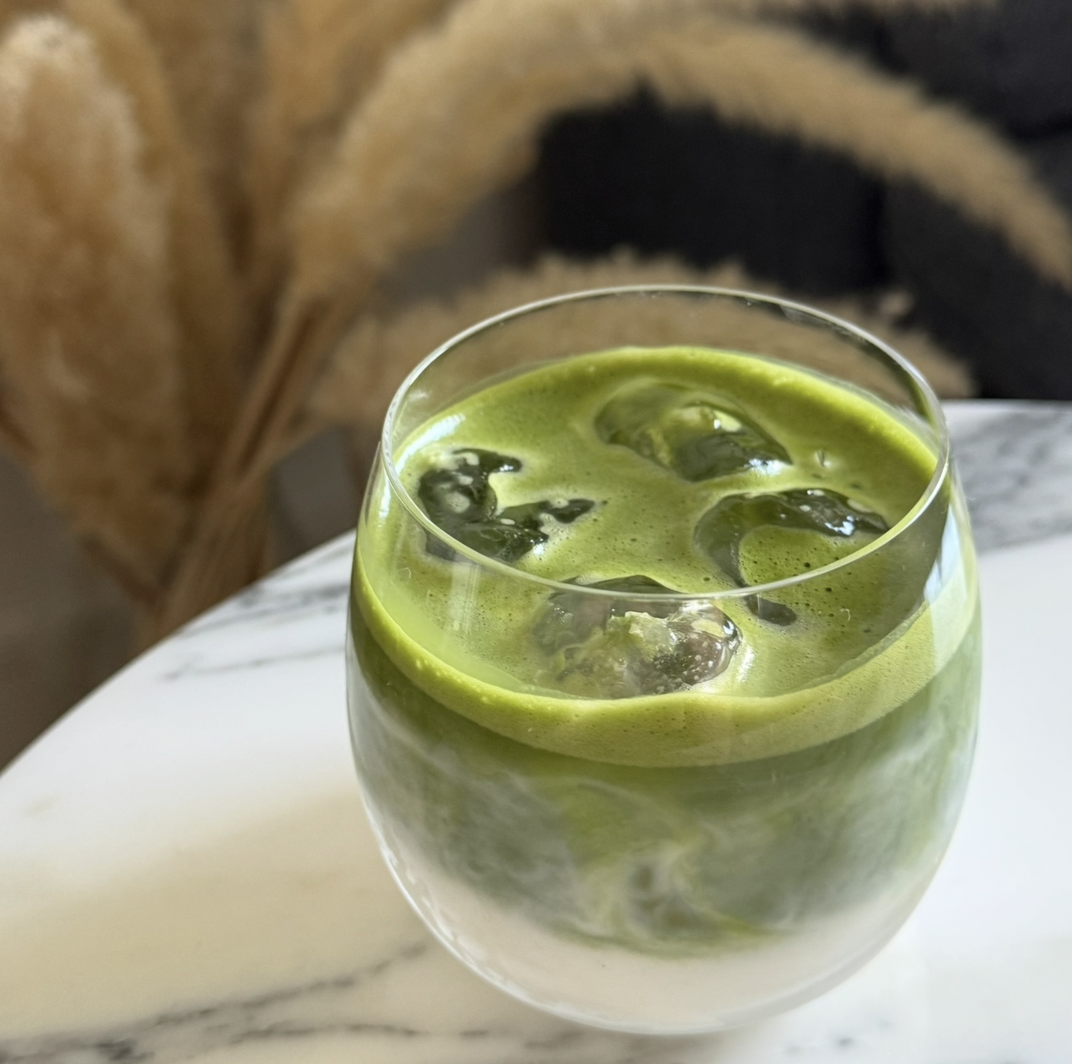 iced matcha latte made using brut tea ceremonial grade matcha powder in the uk, served in a glass tumbler on top of a marble table with decorative pampas grass in the background.