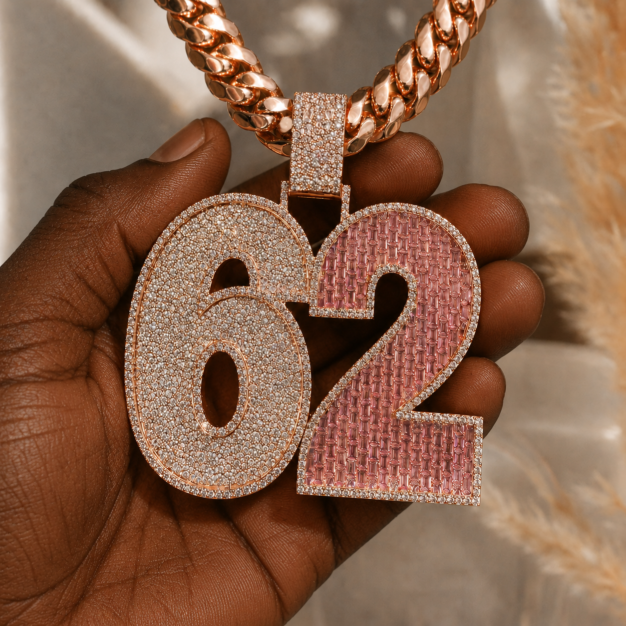 Rose gold iced 62 pendant held by hand, featuring clear pavé stones and pink baguette-style stones.