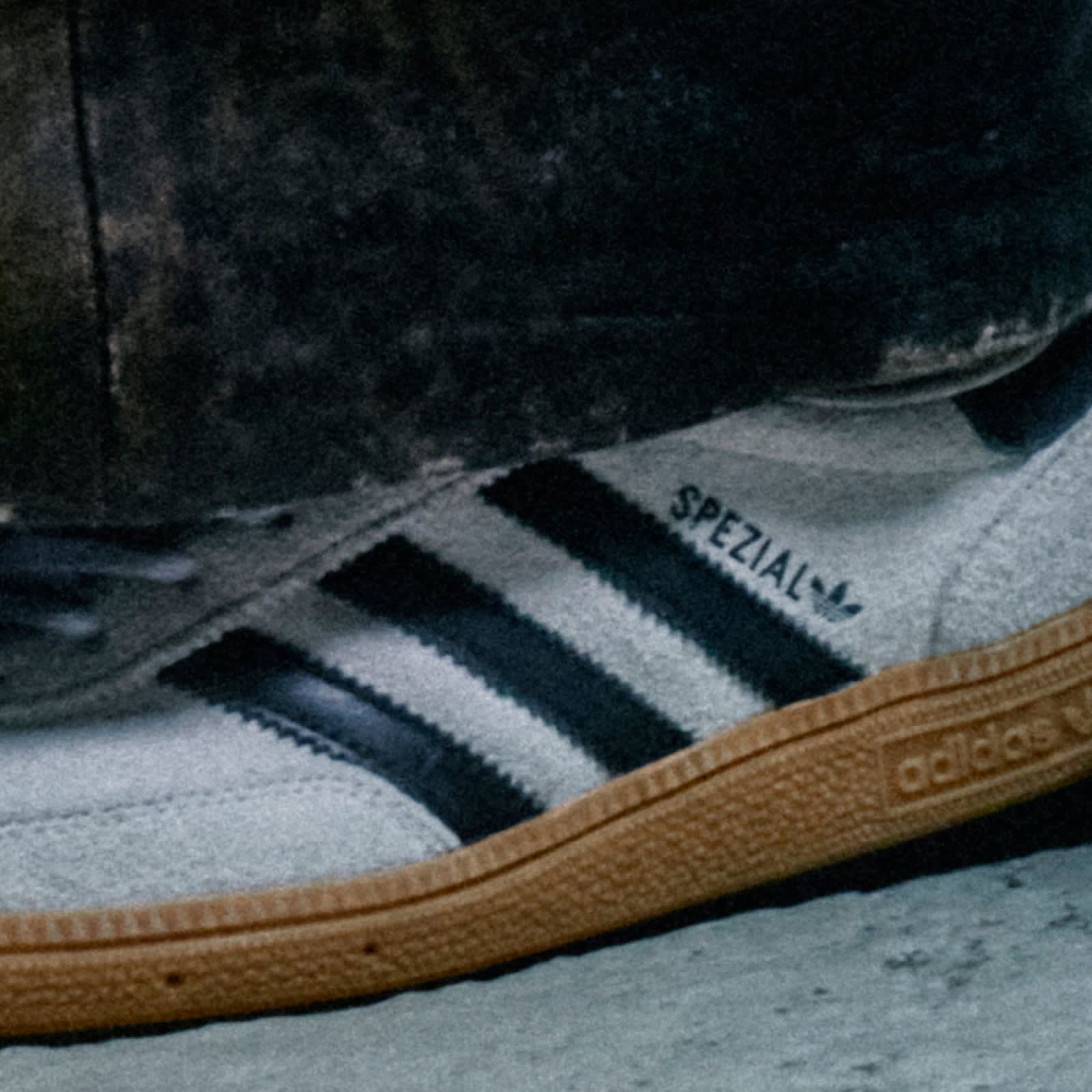 A close-up image of a person wearing a white adidas Spezial sneaker with black stripes and a brown sole standing on a concrete floor. 