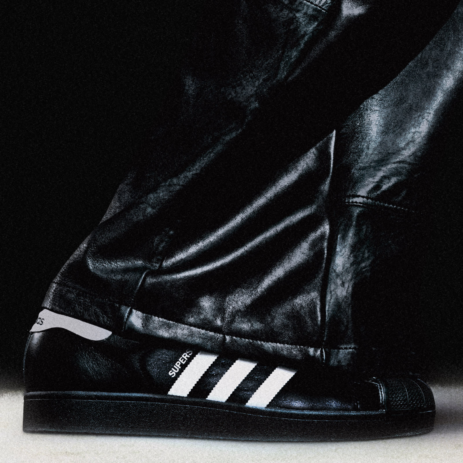 A close-up image of a person wearing a black adidas Superstar sneaker with white stripes and black leather trousers and is standing against a dark background.