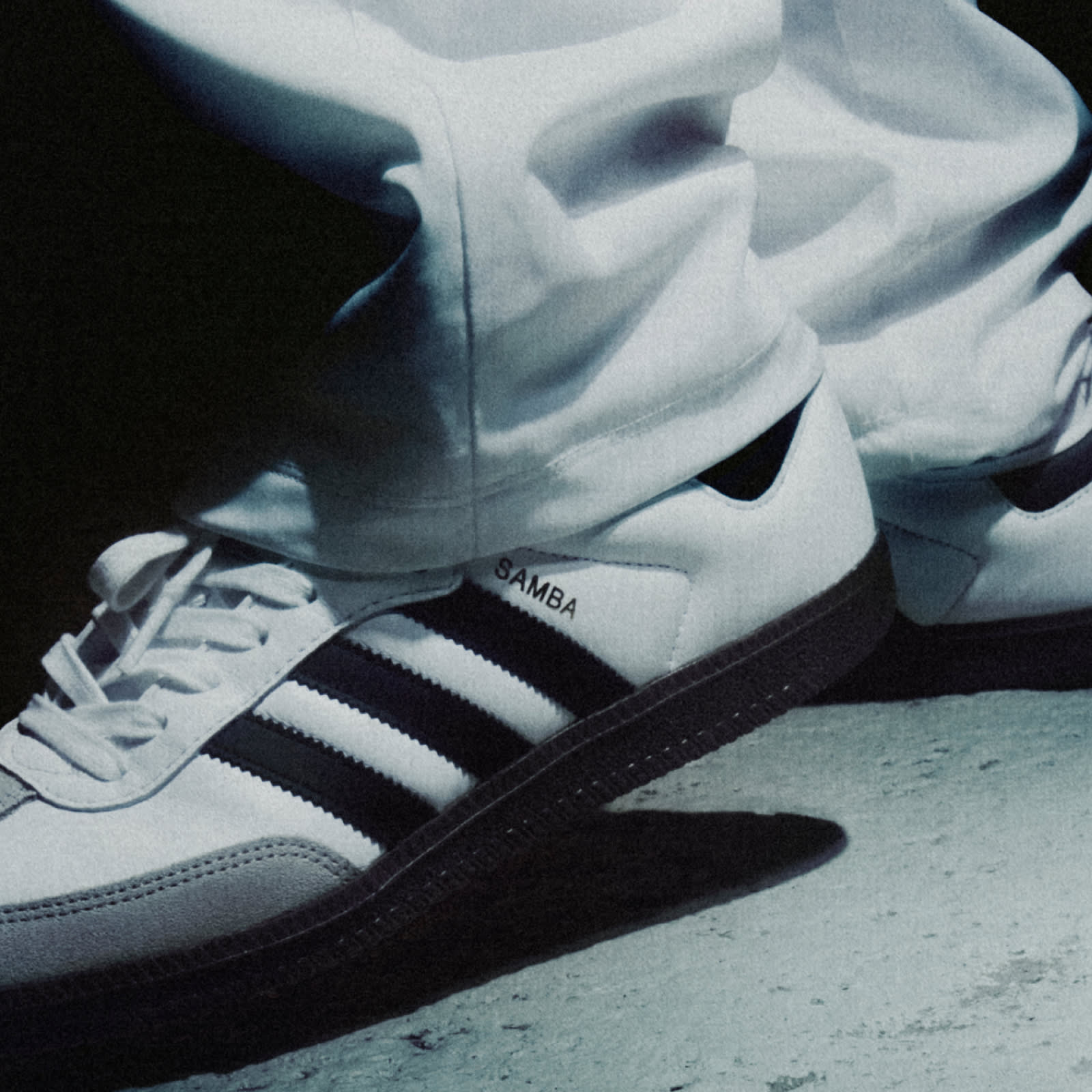 A close-up image of a person wearing a white adidas Samba sneaker with black stripes and white jeans standing on a concrete floor. 