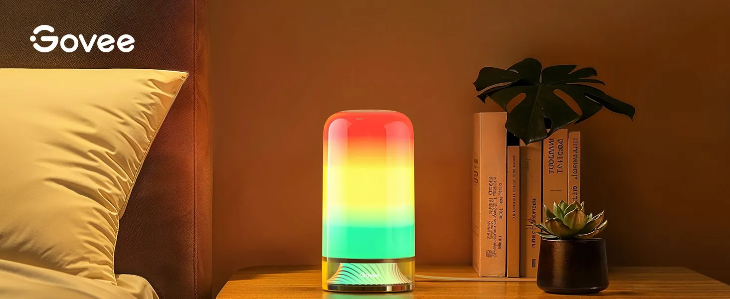 Lampe d'ambiance cylindrique à couleur changeante diffusant une lumière dégradée rouge, jaune et verte, positionnée sur une table de chevet avec des plantes décoratives à proximité