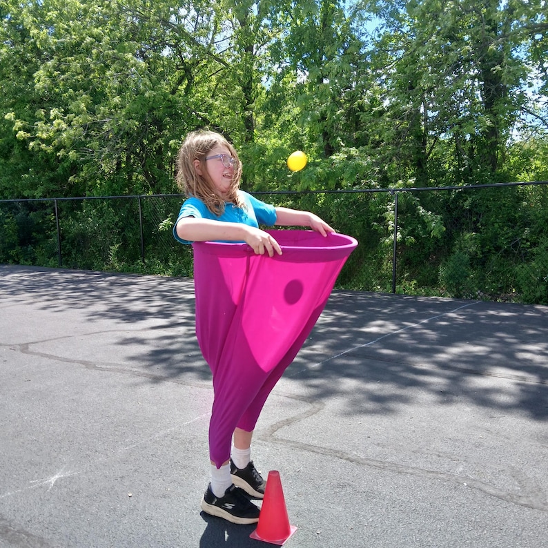 May include: A person holding a pink fabric cone with a hole in the center. A yellow ball is in the air above the cone.