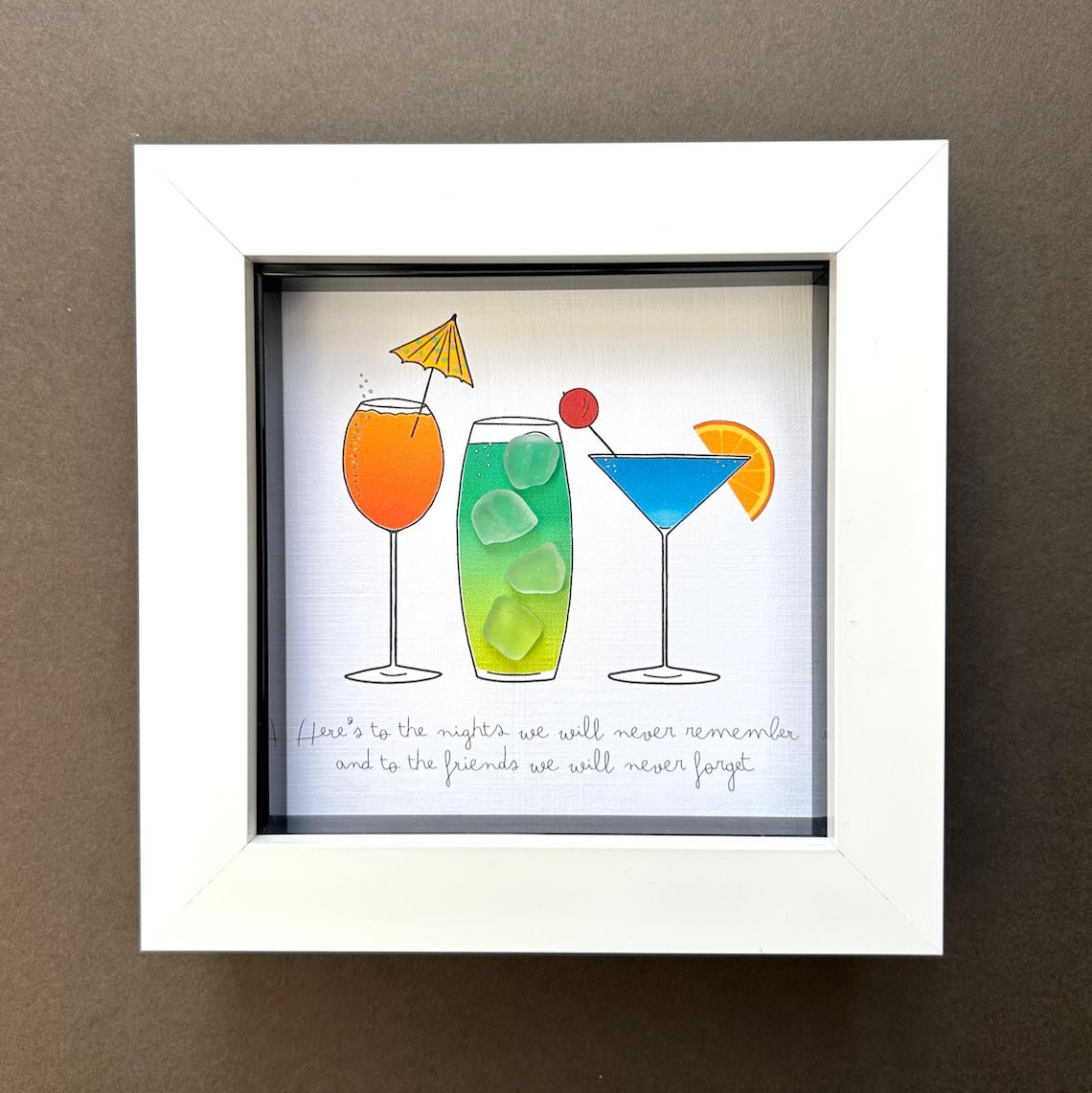 May include: A framed artwork featuring three colorful cocktail illustrations. The drinks are orange, green, and blue, each with unique garnishes like a cherry, orange slice, and a tiny umbrella. The text at the bottom reads, "Here's to the nights we will never remember and to the friends we will never forget."